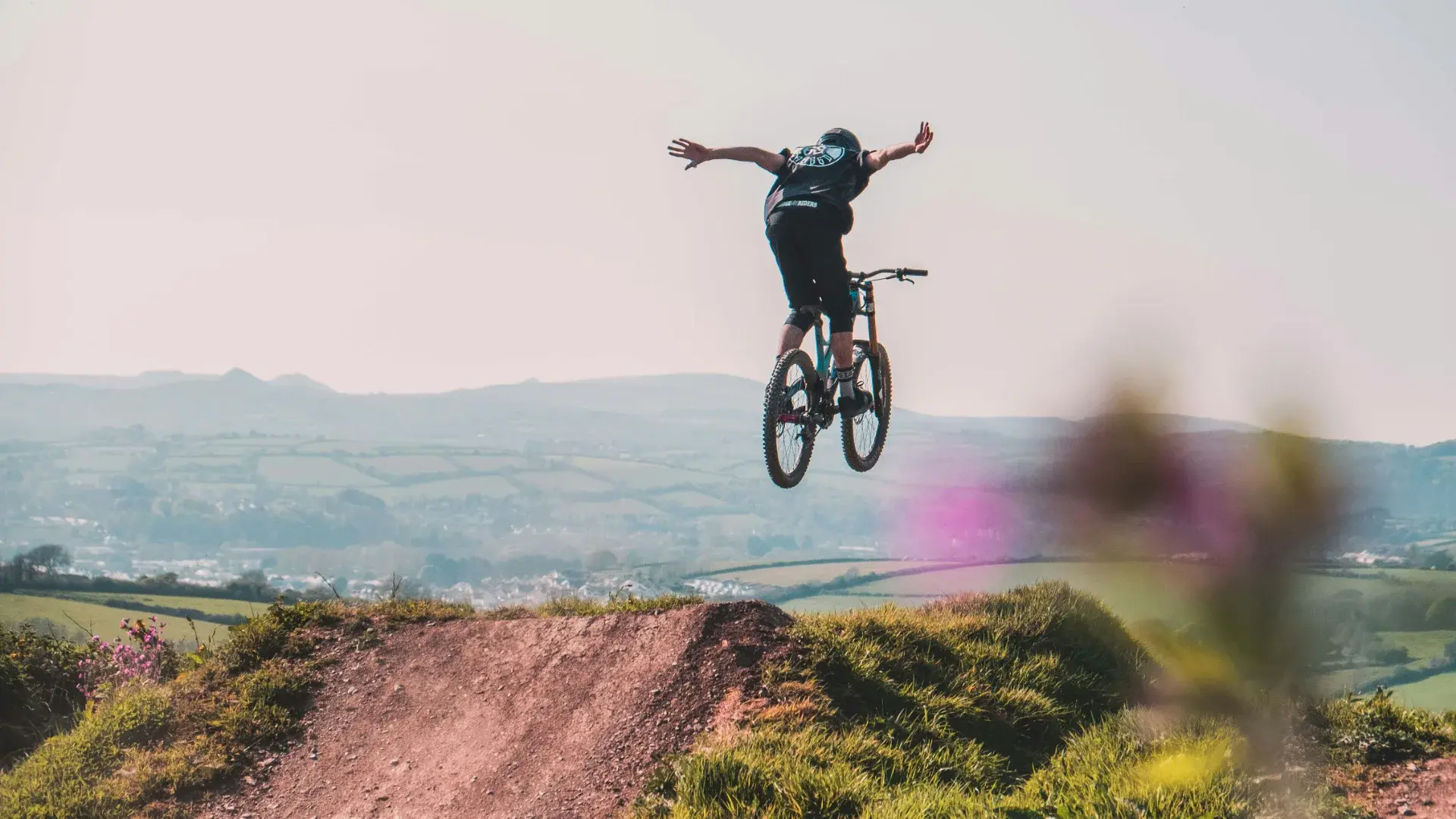Mountainbiker in der Luft über einem großen Table einer Bikepark-Jump-Line, Bergpanorama im Hintergrund